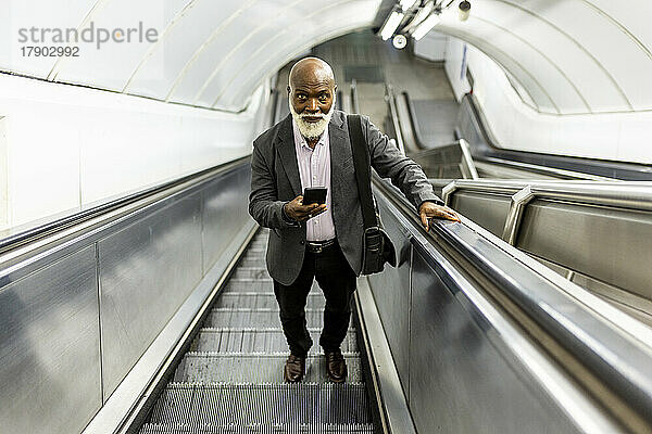 Älterer Pendler mit Glatze und Mobiltelefon bewegt sich auf Rolltreppe nach oben