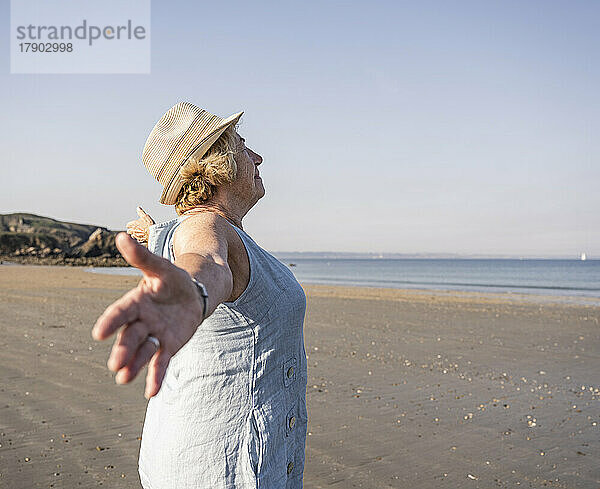 Ältere Frau mit ausgestreckten Armen steht an einem sonnigen Tag am Strand