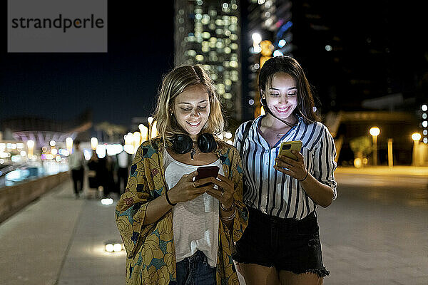 Lächelnde junge Frauen  die nachts in der Stadt Mobiltelefone benutzen