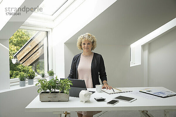 Selbstbewusste blonde Geschäftsfrau steht im Heimbüro am Schreibtisch