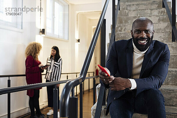 Lächelnder Geschäftsmann mit Smartphone sitzt auf der Treppe  Kollegen unterhalten sich im Hintergrund