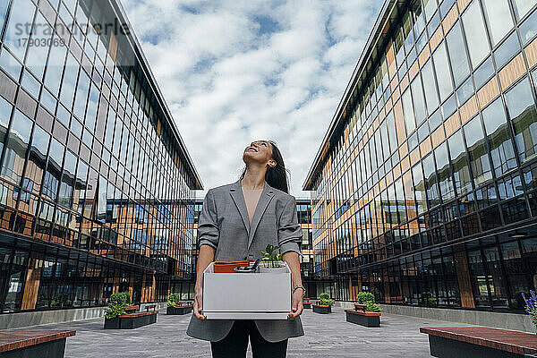 Lächelnde Geschäftsfrau mit Karton vor Bürogebäude unter bewölktem Himmel