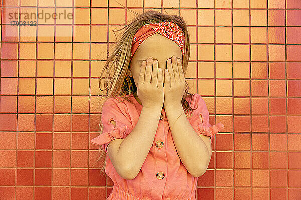 Mädchen bedeckt Gesicht mit Händen und steht vor der Wand