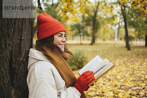 Glückliche junge Frau hält Buch am Baumstamm im Park