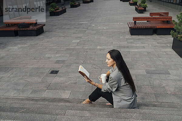 Geschäftsfrau mit Kaffeetasse liest Buch auf Stufen im Büropark