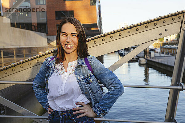 Glückliche Frau mit den Händen auf der Hüfte steht auf einer Brücke über den Fluss  Hafencity  Hamburg  Deutschland