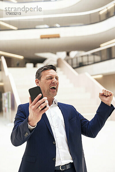 Fröhlicher Geschäftsmann mit Smartphone und geballter Faust in der Lobby