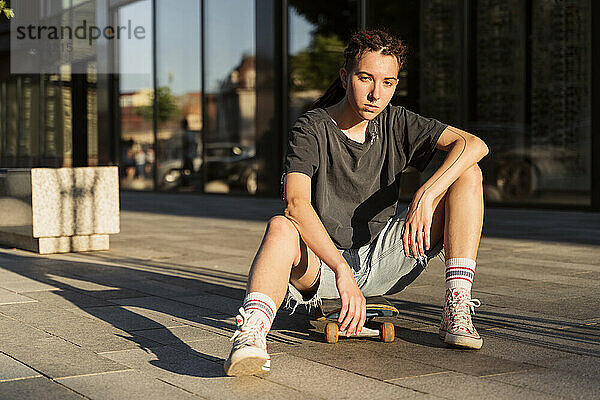 Junge nicht-binäre Person sitzt auf einem Skateboard am Fußweg