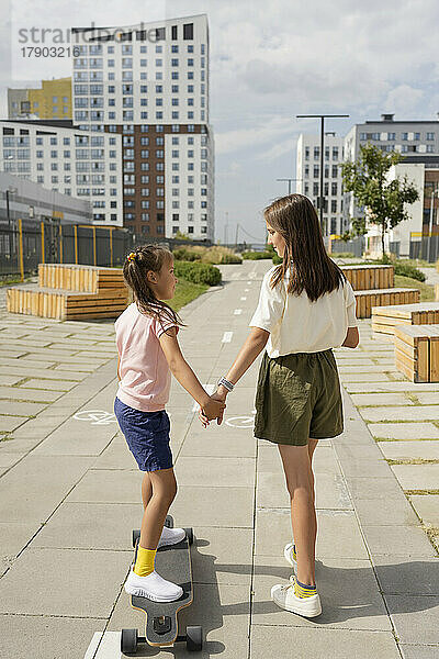 Mädchen hält die Hand ihrer Schwester und unterrichtet an einem sonnigen Tag auf dem Fußweg Longboard-Skaten