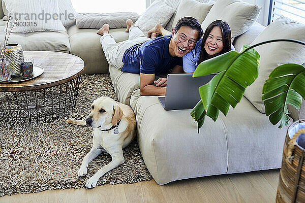 Fröhliches Paar mit Laptop  das zu Hause auf dem Sofa im Wohnzimmer liegt