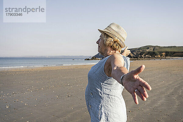 Ältere Frau mit ausgestreckten Armen steht am Strand