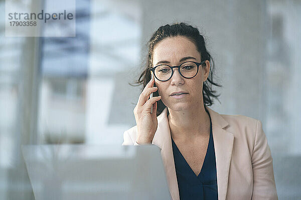 Geschäftsfrau telefoniert mit Laptop und sitzt durch Glas vor einer grauen Wand