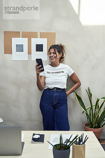 Lächelnde Geschäftsfrau benutzt Smartphone und lehnt an der Wand