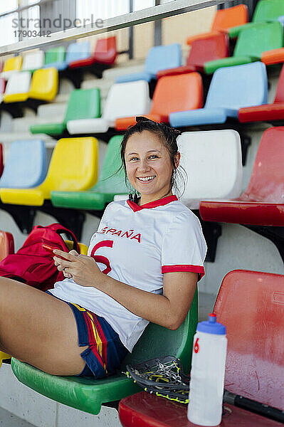 Happy player sitting on seat with smart phone at stadium