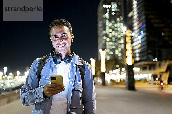Glücklicher Mann mit kabellosen Kopfhörern  der nachts sein Mobiltelefon in der Hand hält