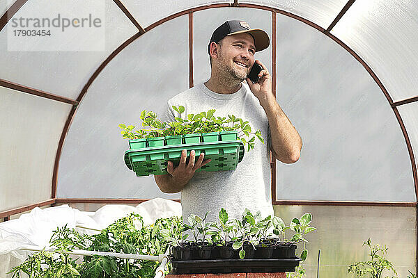 Glücklicher Bauer hält Topfpflanzen in der Hand und telefoniert im Gewächshaus