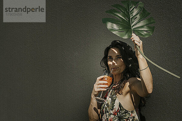 Woman having drink holding monstera leaf in front of wall
