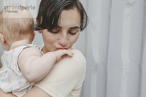 Mother kissing baby girl's little hand in front of wall