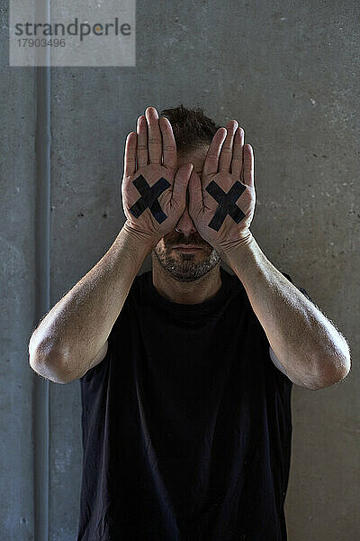 Mann bedeckt die Augen mit den Händen und zeigt ein Kreuz vor der Wand