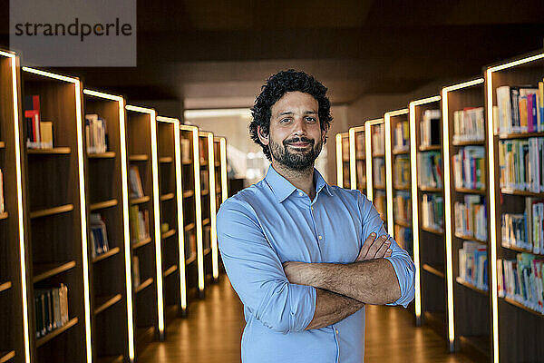 Lächelnder Mann mit verschränkten Armen inmitten von Bücherregalen in beleuchteter Bibliothek