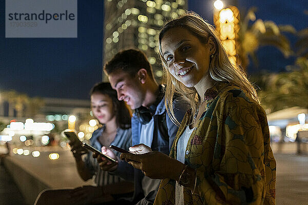 Lächelnde Frau mit Mobiltelefon  die nachts bei Freunden sitzt