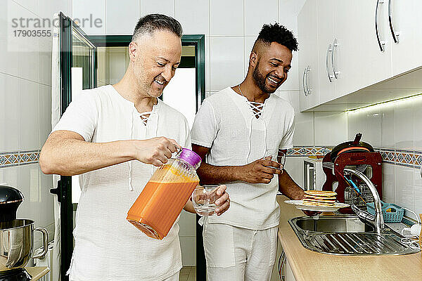 Glücklicher reifer Mann gießt Saft in Glas  während sein Freund einen Teller Pfannkuchen in der Küche hält