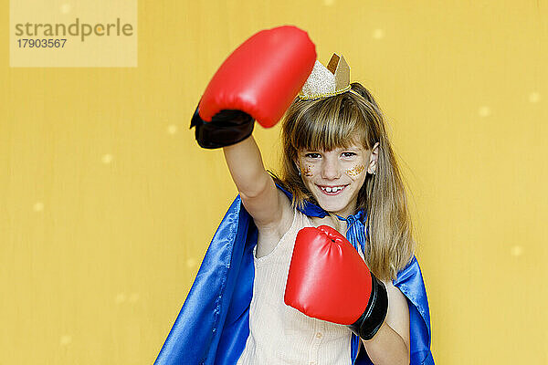 Happy girl wearing boxing gloves and cape against yellow background