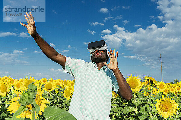 Lächelnder junger Mann mit Virtual-Reality-Simulator  der im Sonnenblumenfeld tanzt