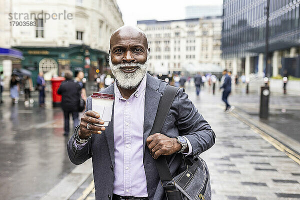 Lächelnder älterer Geschäftsmann mit Laptoptasche beim Kaffeetrinken auf dem Fußweg