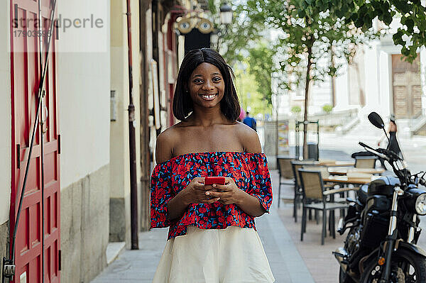 Glückliche junge Frau mit Smartphone steht auf Fußweg