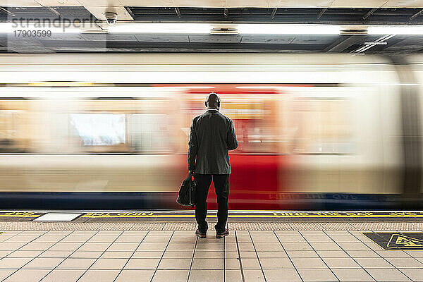 Älterer Passagier mit Laptoptasche steht vor einem rasenden Zug auf dem U-Bahnsteig