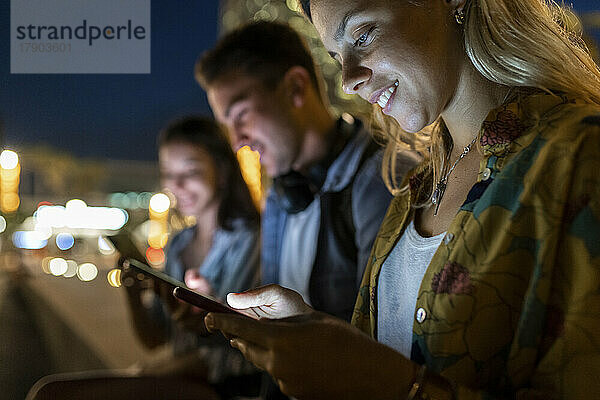 Lächelnde junge Frau  die nachts ihr Mobiltelefon benutzt und bei Freunden sitzt