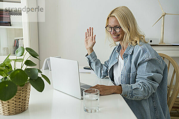 Lächelnder Freiberufler winkt bei einem Videoanruf über den Laptop zu Hause mit der Hand