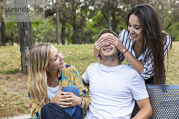 Glückliche Frau bedeckt die Augen eines Mannes  der neben einem Freund auf einer Bank im Park sitzt