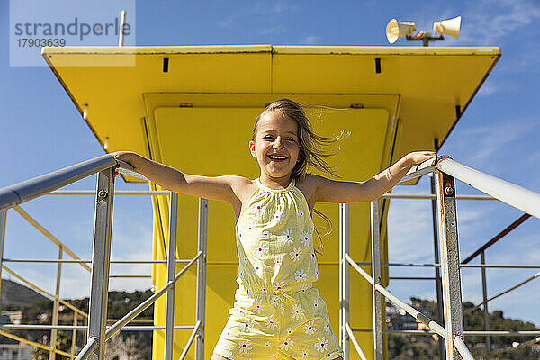 Fröhliches Mädchen  das an einem sonnigen Tag vor der gelben Rettungsschwimmerhütte steht