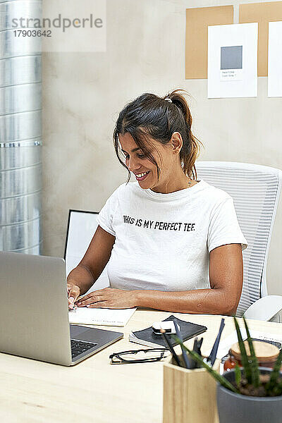 Lächelnde Geschäftsfrau schreibt im Notizblock und arbeitet im Heimbüro