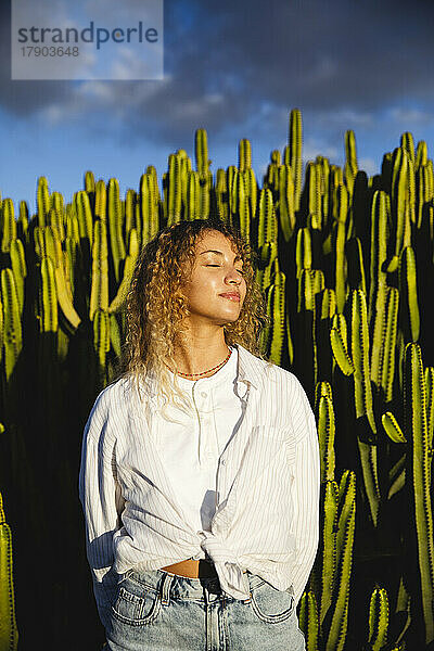 Young woman with eyes closed enjoying sunlight in front of cactus