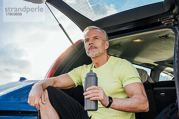 Reifer Mann mit Flasche sitzt im Kofferraum eines Autos