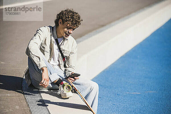 Glücklicher Mann mit Skateboard und Smartphone am Rande der Leichtathletik