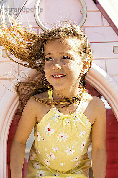 Glückliches Mädchen mit zerzausten Haaren  das an einem sonnigen Tag durch das Fenster des Spielhauses schaut