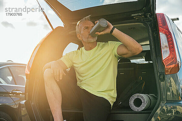 Reifer Mann trinkt Wasser im Kofferraum eines Autos