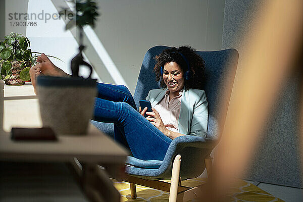Glückliche Geschäftsfrau  die am Arbeitsplatz auf einem Stuhl sitzt und ihr Mobiltelefon benutzt