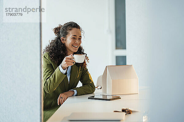 Glückliche Geschäftsfrau mit Kaffeetasse und Modellhaus auf dem Schreibtisch im Büro