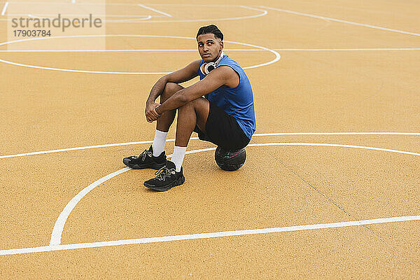 Nachdenklicher junger Mann sitzt auf Basketball auf dem Sportplatz