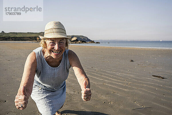 Glückliche ältere Frau zeigt an einem sonnigen Tag am Strand Daumen nach oben