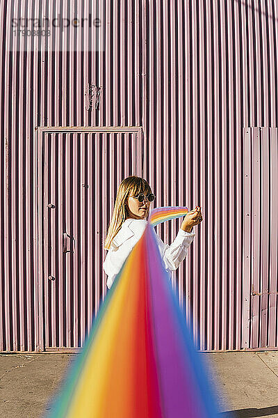 Junge Frau blickt über die Schulter und hält ein regenbogenfarbenes Band vor einer Wellwand