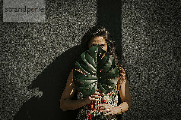 Reife Frau bedeckt Gesicht mit Monstera-Blatt vor der Wand