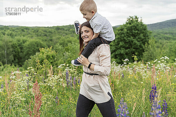 Lächelnde junge Frau trägt Sohn auf Schultern auf der Wiese