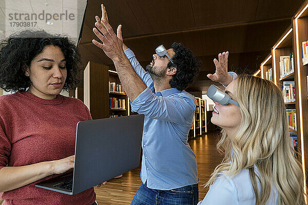 Frau arbeitet am Laptop  während Kollegen mit VR-Brille in der Bibliothek gestikulieren