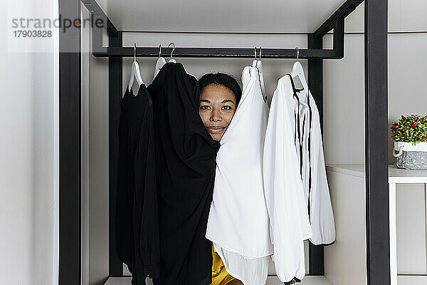 Woman sitting inside closet and peeking out from behind clothes